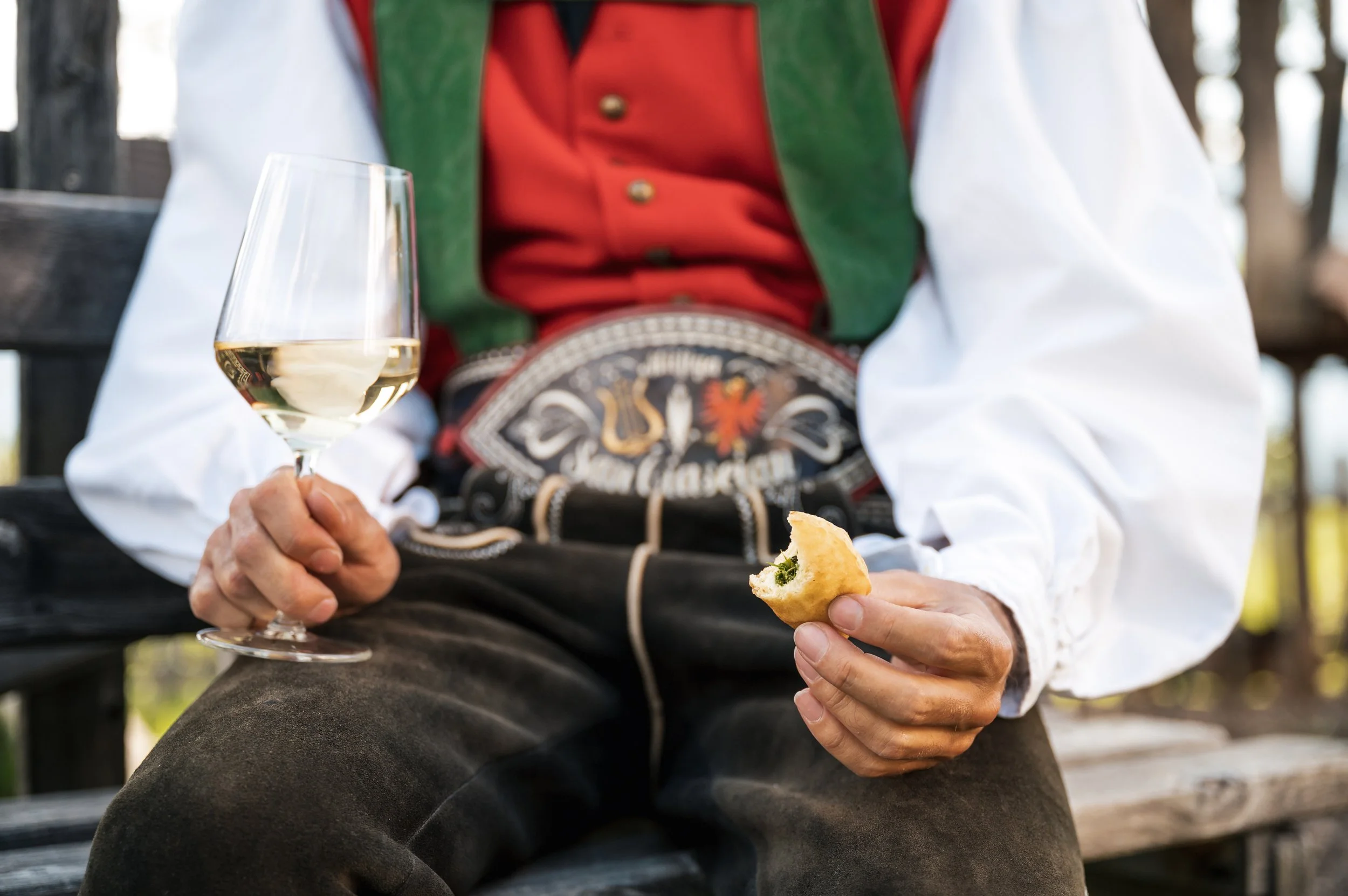 Person in traditional German attire sitting on a bench, holding a glass of white wine in one hand and a partially eaten pretzel in the other.