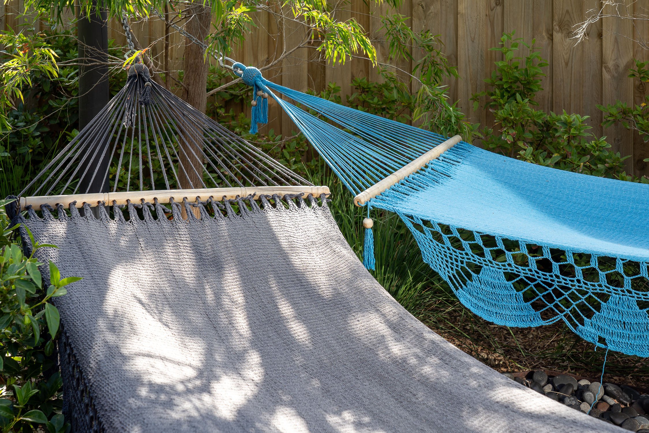 Two hammocks, one grey and one blue, hanging side by side outdoors with a wooden fence and greenery in the background.