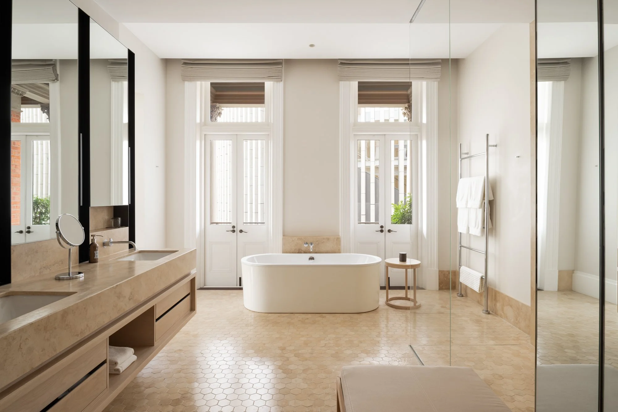 Modern bathroom with a freestanding bathtub, large windows with blinds, a double sink vanity with a mirror, a towel rack, and neutral-toned tile flooring.