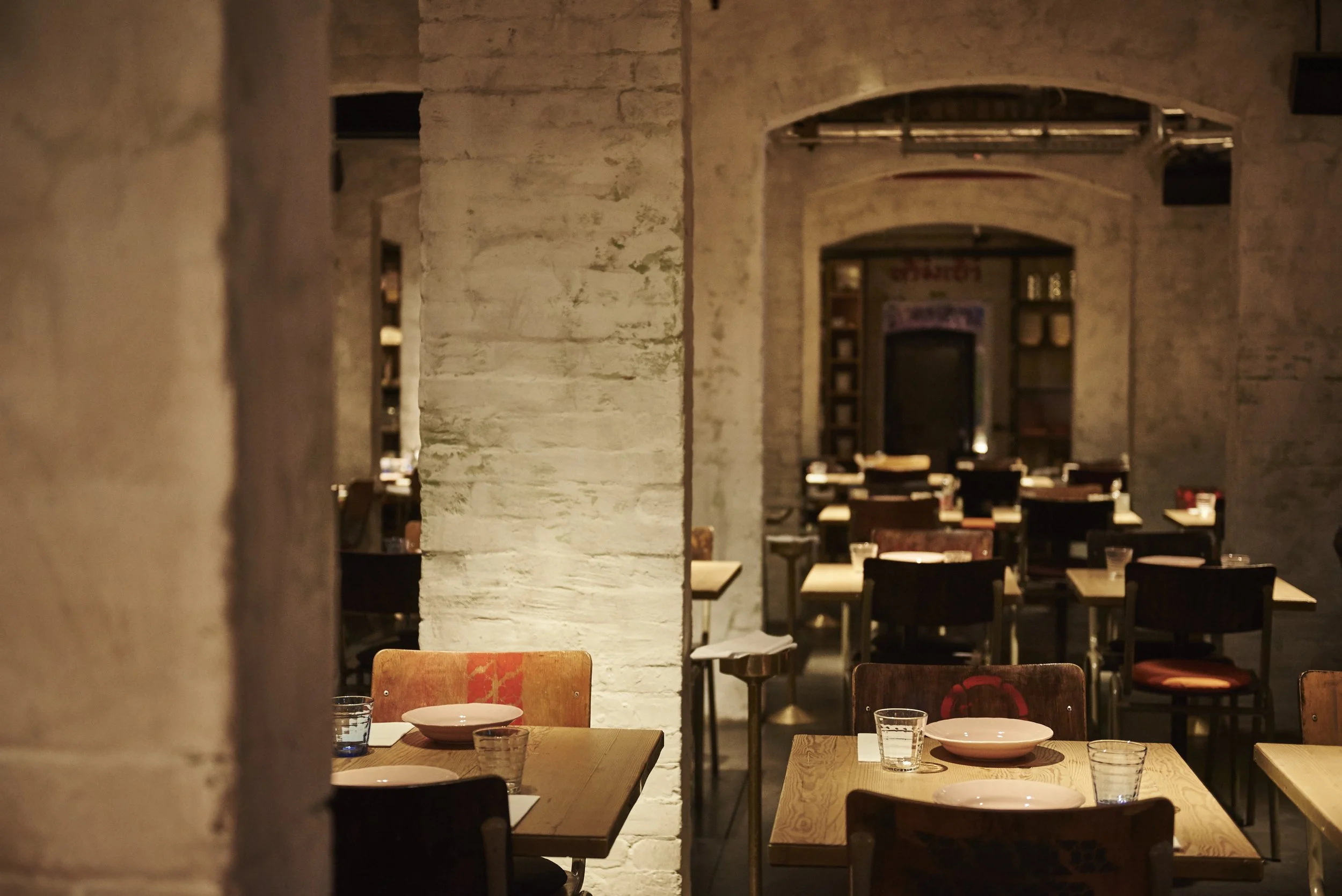 Empty restaurant with wooden tables, chairs, white plates, and drinking glasses, illuminated by warm lighting and brick walls.