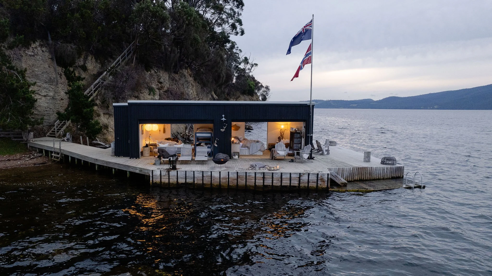 A houseboat on calm water with an outdoor deck, illuminated interior, and flags flying on a flagpole. Hills and overcast sky in the background.