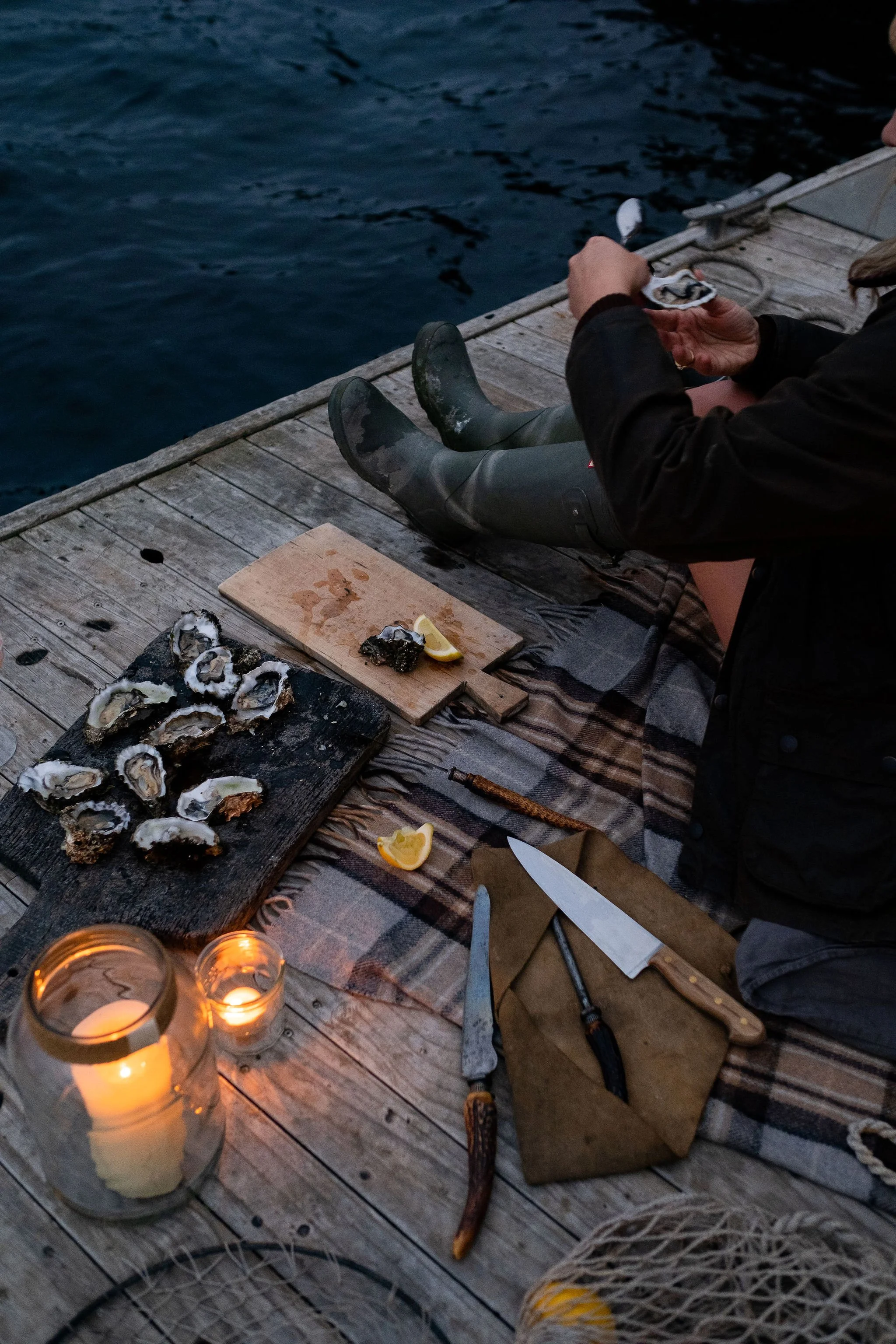A person sitting on a wooden dock by the water, opening a can or a container. There is a cutting board with oysters, lemon wedges, and a black object. A blanket is spread on the dock. Two candles are lit inside jars, providing warm light. There are knives on a brown cloth, and some oysters on a black platter. The person is wearing rubber boots, dark pants, and a coat.