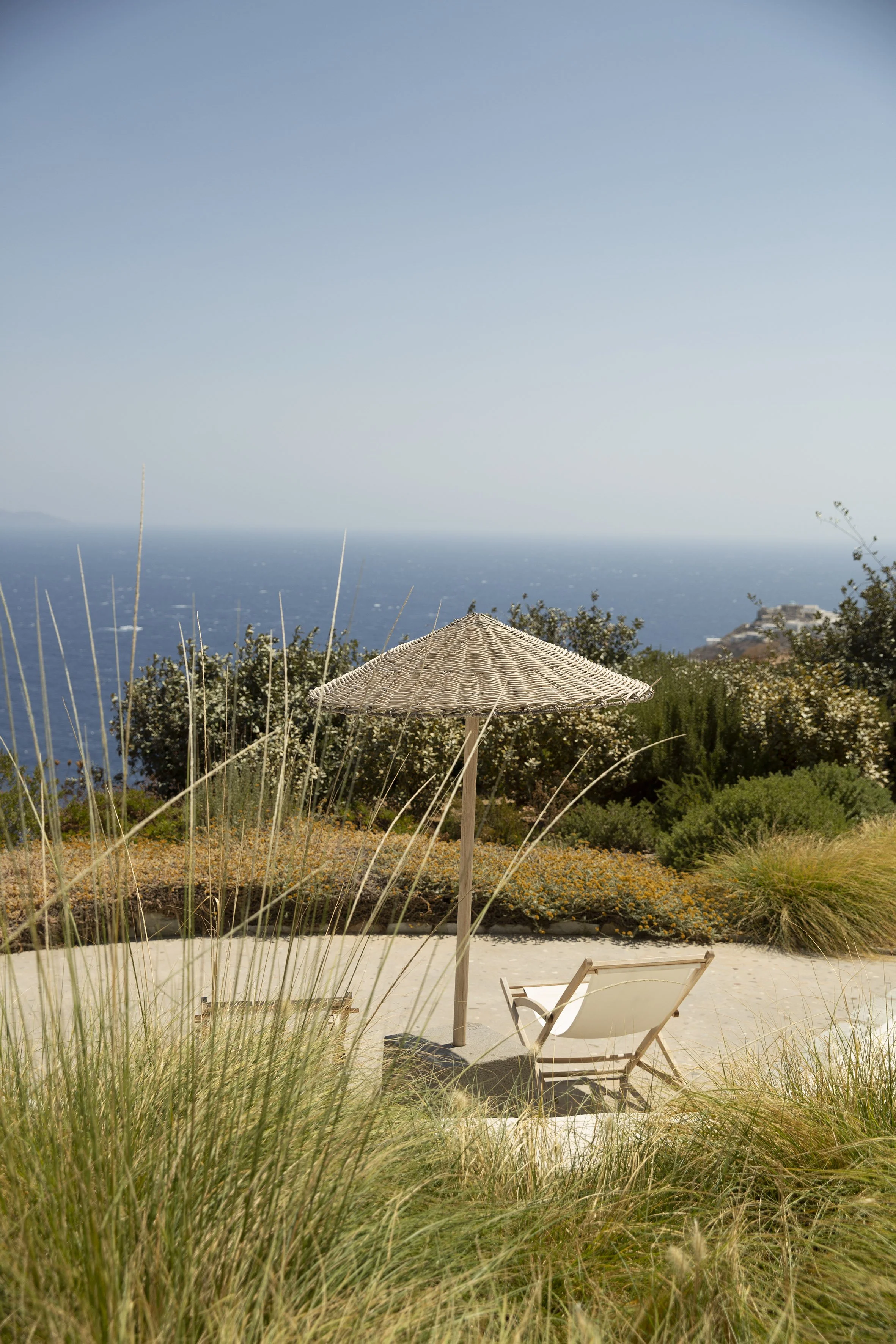 Beach scene with a beach chair, a woven umbrella, surrounded by tall grass and shrubs, overlooking the ocean under a clear blue sky.