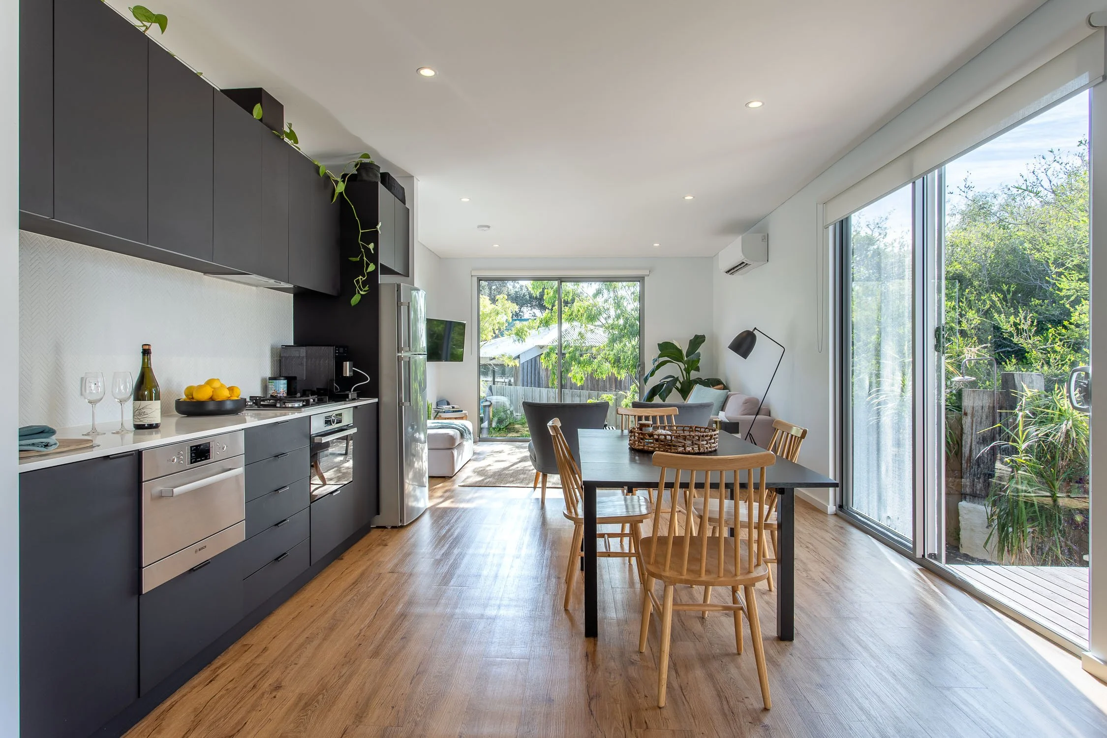 Open-concept living and dining area in a modern home with hardwood floors, gray kitchen cabinets, a dining table with wooden chairs, and large sliding glass doors leading to an outdoor space with greenery.