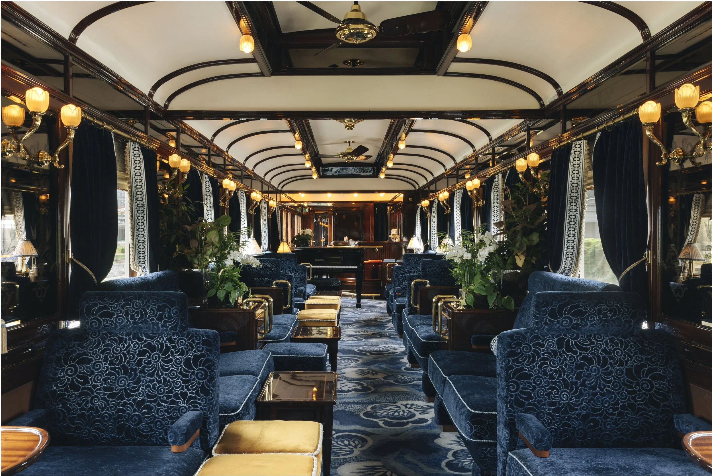 Interior of a vintage train carriage with elegant blue velvet armchairs, small tables, and potted plants. Warm lighting along the walls and decorative curtains adorn the windows, with a piano at the far end.