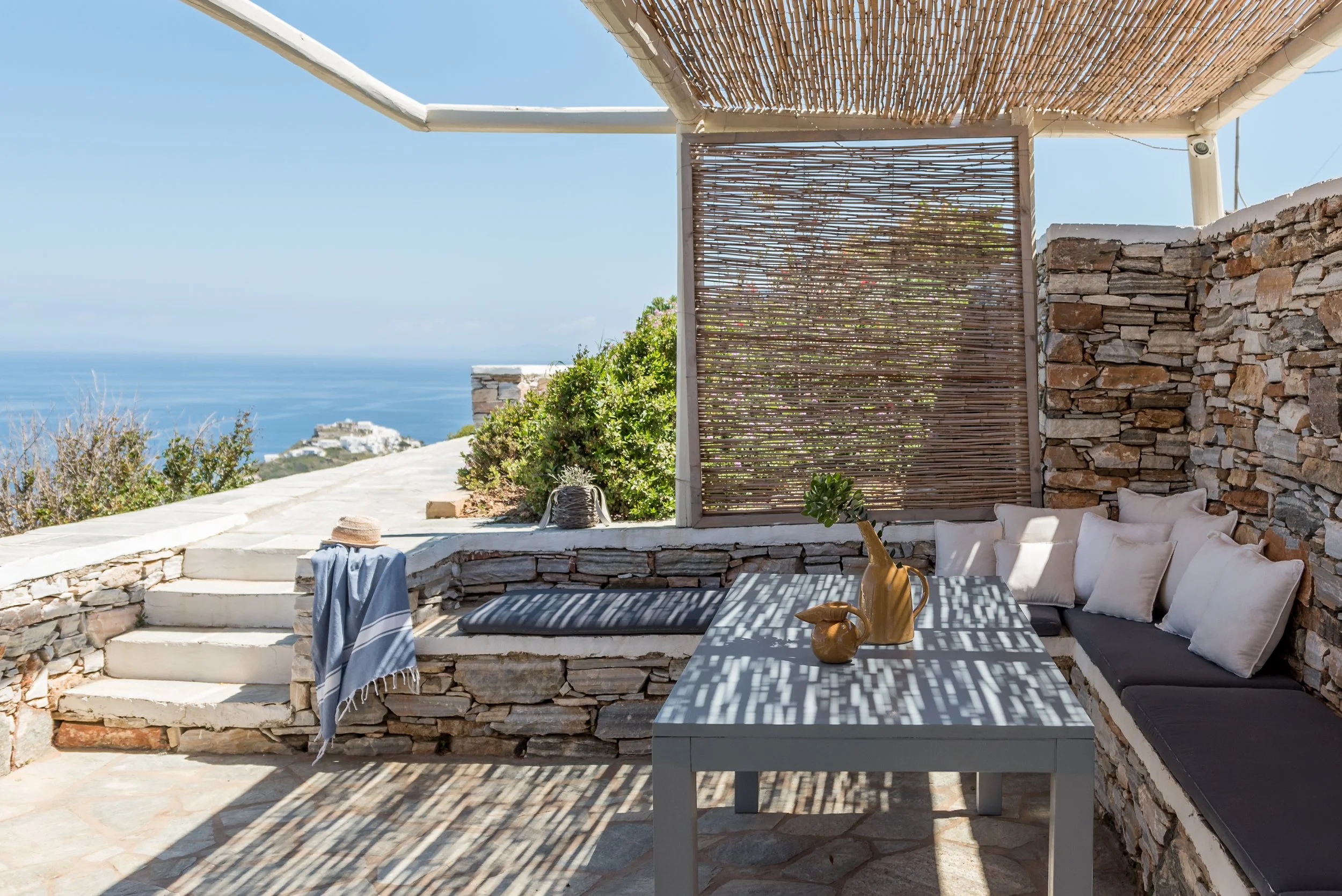 Outdoor patio with stone walls, a black cushioned bench with white pillows, a rectangular table with vases, and a view of the ocean and sky with greenery.
