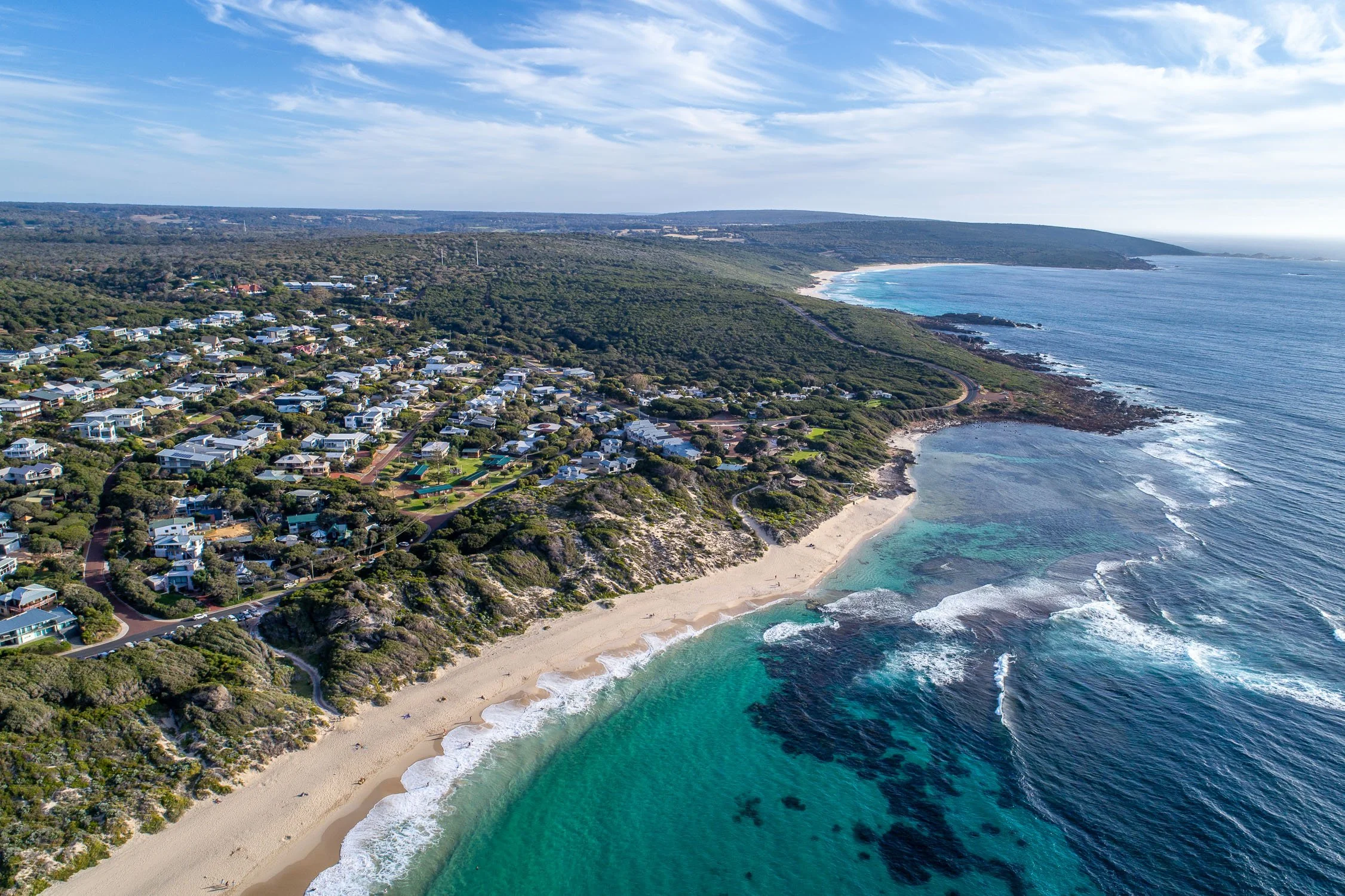 Aerial view of a coastal town with numerous white houses, green vegetation, rocky cliffs, sandy beaches, and the ocean with waves and coral reefs under a partly cloudy sky.
