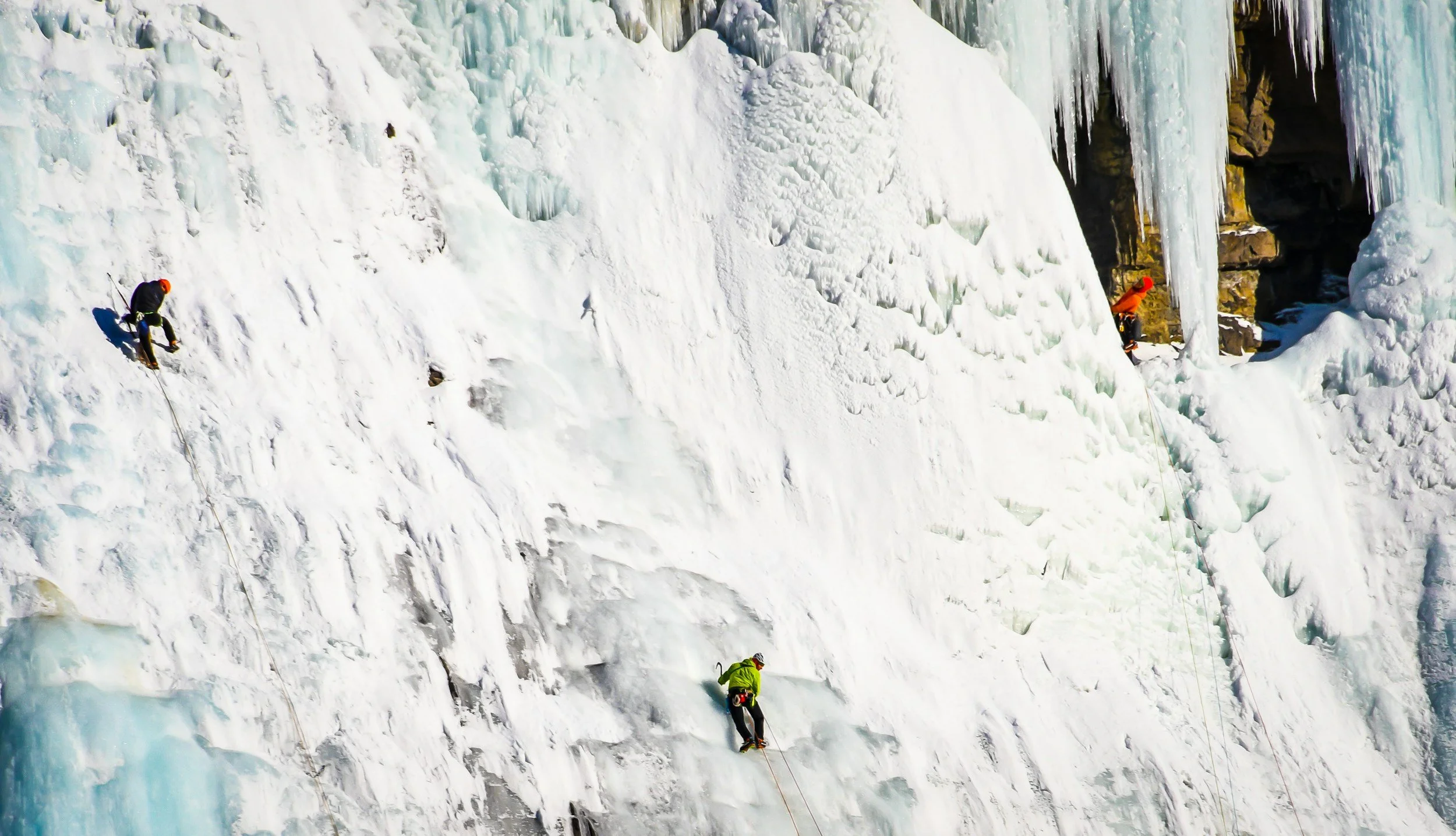 Three ice climbers ascending a vertical ice wall in a snowy and icy landscape, with ice formations and icicles surrounding them.