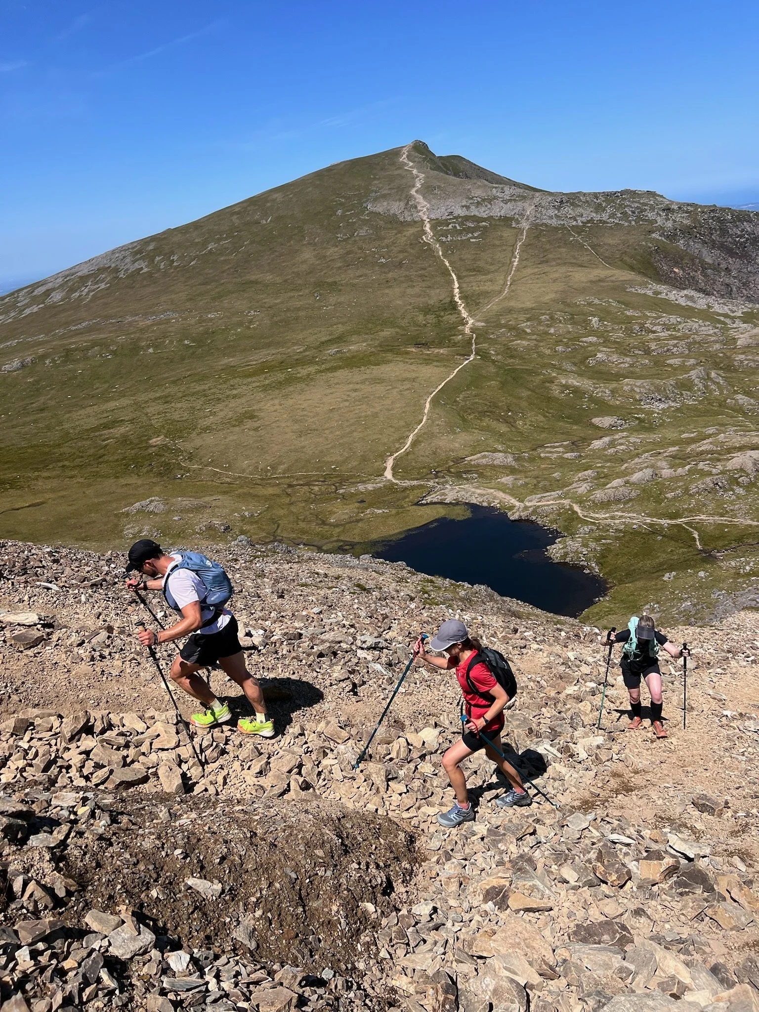 Three hikers ascending a rocky mountain trail with a grassy hill, a small lake, and a clear blue sky in the background.