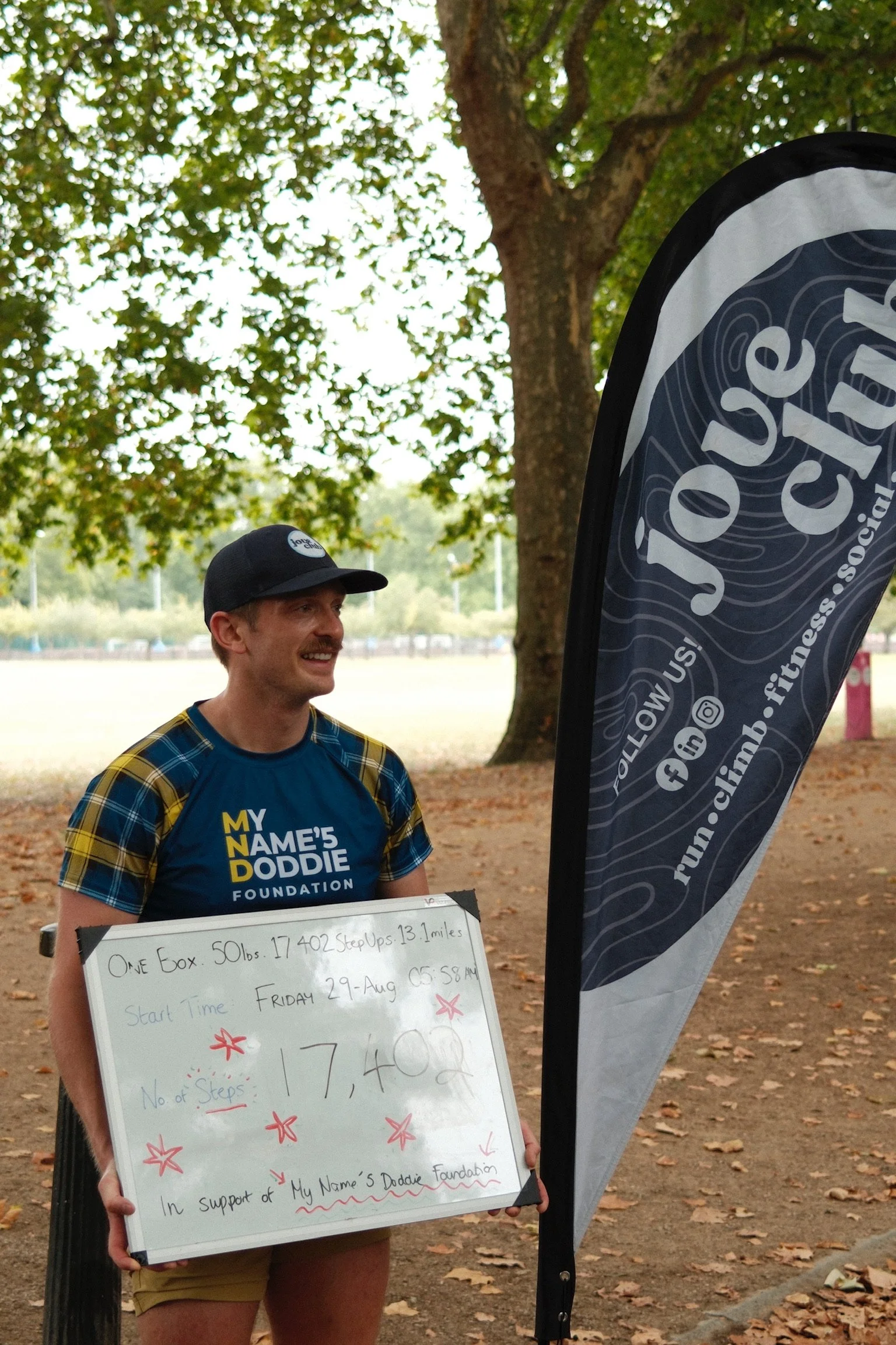 A man participating in a running event, holding a whiteboard with his race details, standing outdoors near a tree and a promotional flag for 'run.cim.fitness.social'.