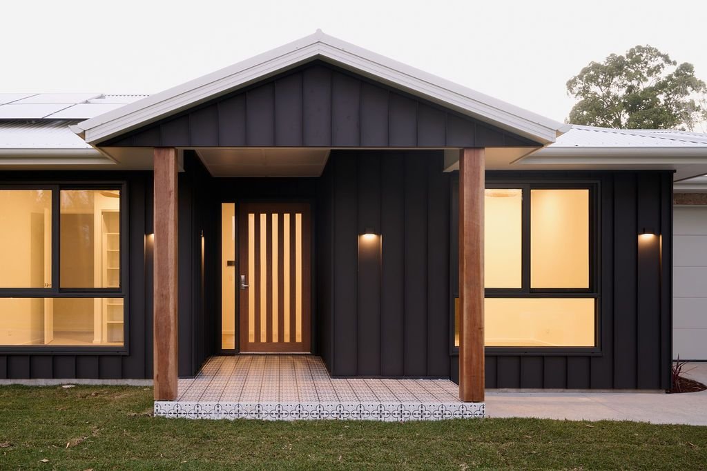 Front view of a modern house with black exterior walls, large windows, wooden posts, and a small porch with patterned tile flooring, illuminated by exterior lights at dusk.