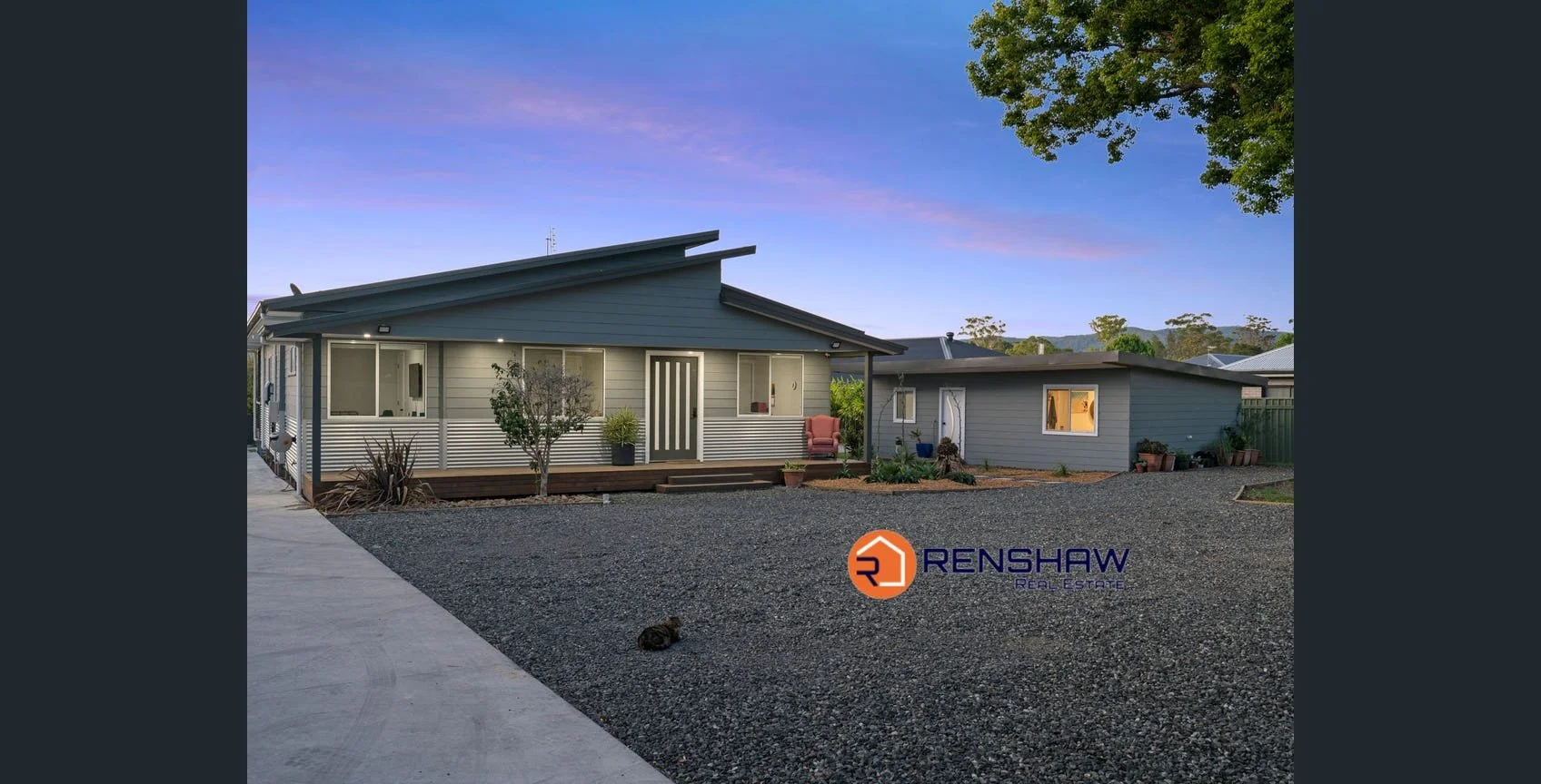 Photo of a modern, single-story house with a grey exterior and a sloped roof at dusk. The front yard has gravel, some plants, and a small tree. There is a porch with seating and potted plants. The Renshaw Real Estate logo is in the bottom right corner.