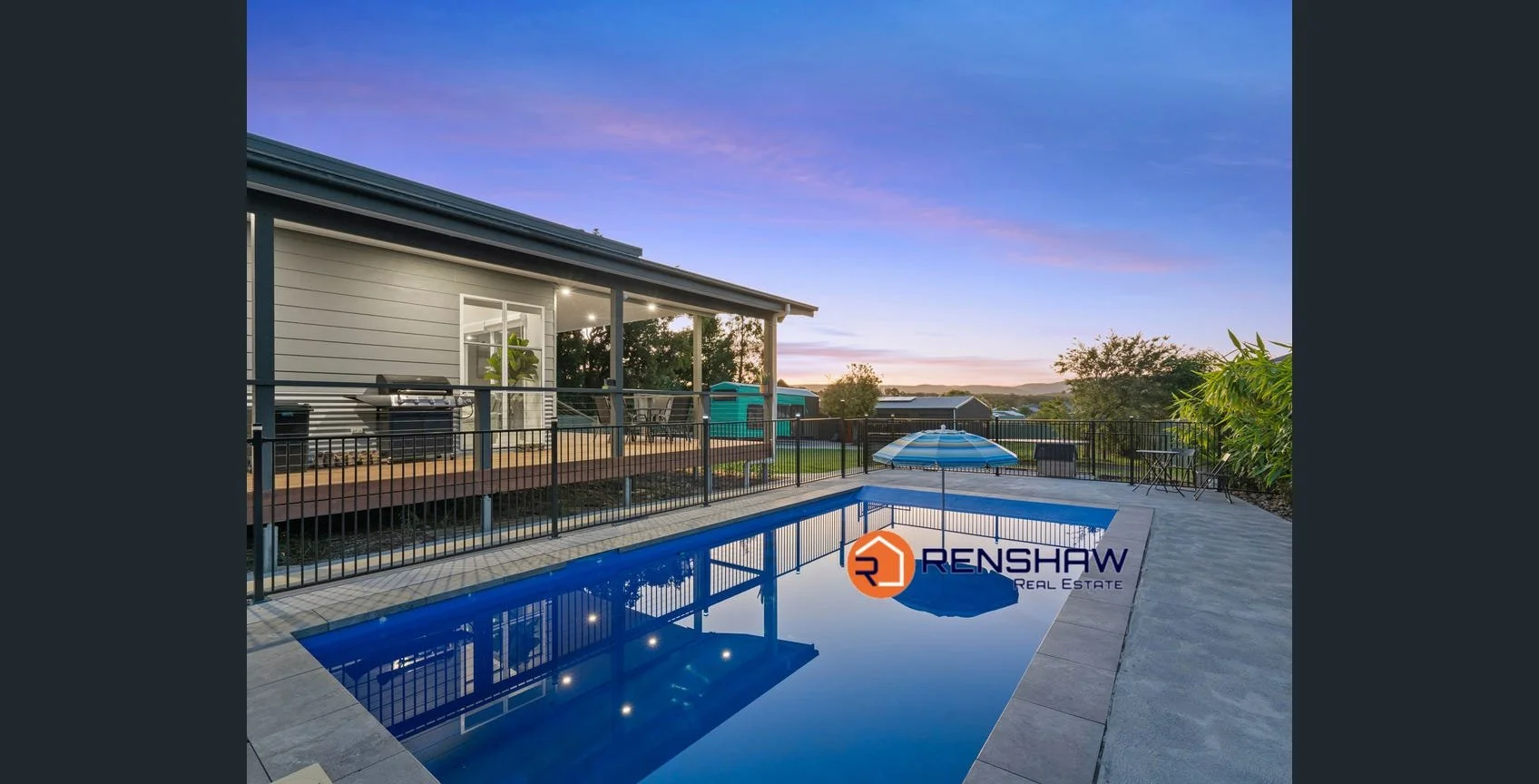 Residential backyard with a swimming pool, patio with table and umbrella, deck with grill, fence, and surrounding trees during sunset.