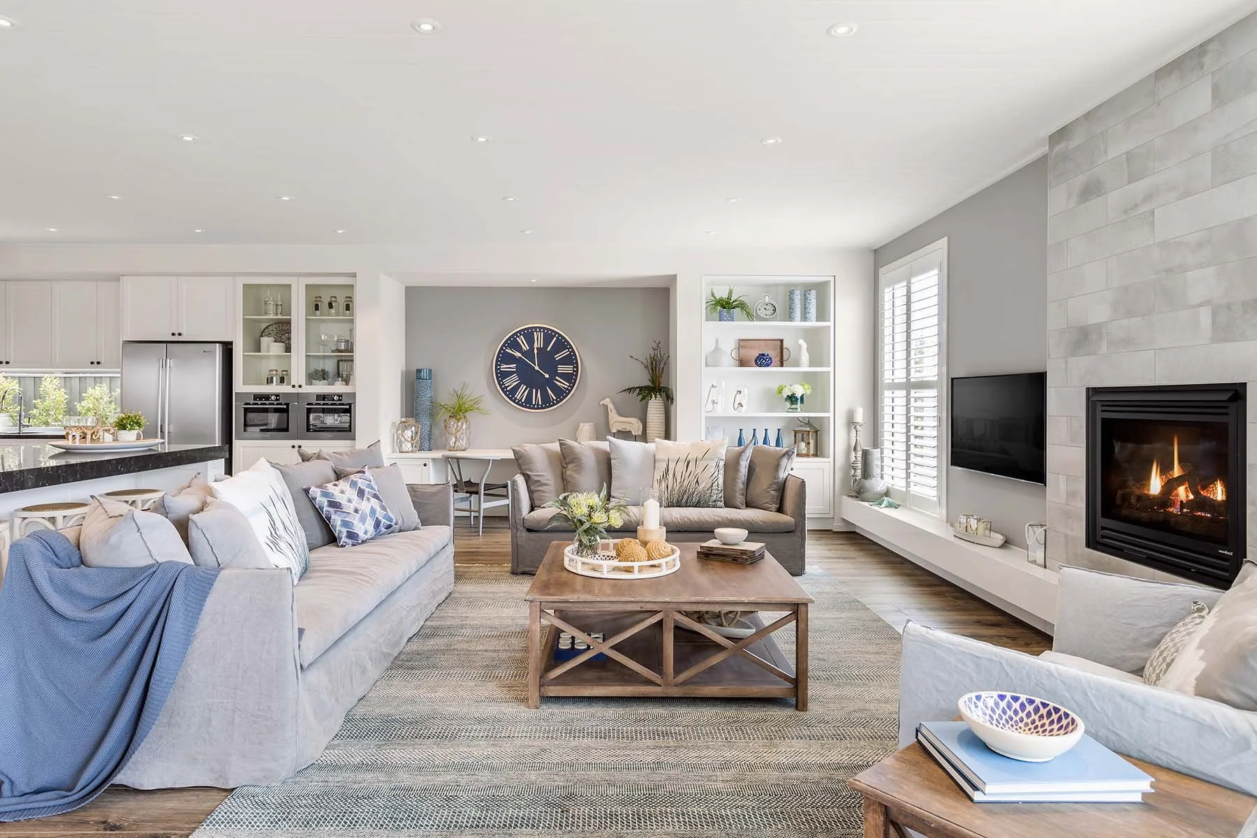 Bright, modern living room with gray and white tones, a fireplace on the right, a large flat-screen TV, and a wooden coffee table in the center decorated with candles and flowers.