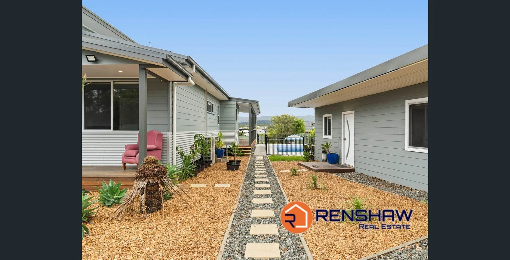 View of a backyard with a pathway of stepping stones flanked by gravel and small plants, between two houses with light blue siding, a pink armchair on the porch of the left house, and a swimming pool and trees in the background. Renshaw Real Estate logo at the bottom right.