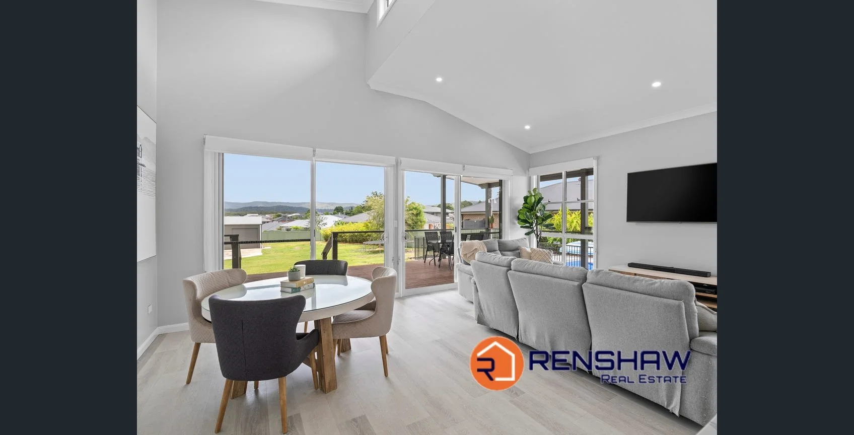 Living room with large windows, gray sofas, a wall-mounted TV, a round dining table with four chairs, and a view of a backyard with a deck, lawn, and distant hills.