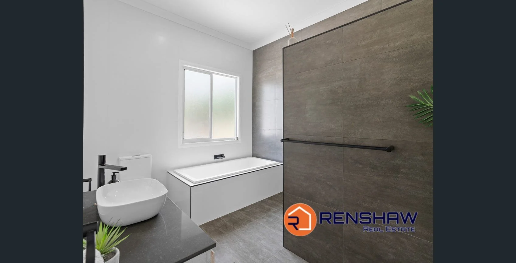 Modern bathroom with a white bathtub beneath a frosted window, a white vessel sink on a dark countertop, and a large tile walk-in shower with a black grab bar. The Renshaw Real Estate logo is in the bottom right corner.