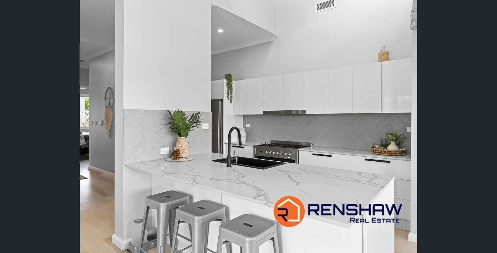 Modern white kitchen with marble countertop, black sink, and minimalist cabinets. Decor includes green plants and woven accessories. Renshaw Real Estate logo in the bottom right corner.