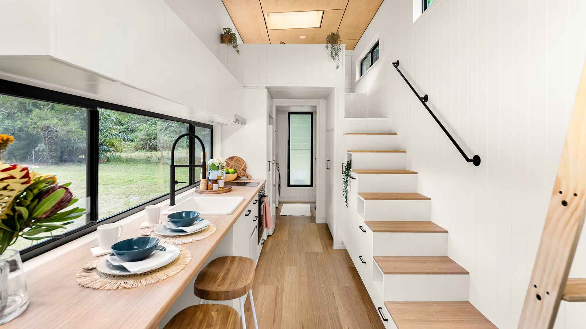 Bright, modern kitchen with a large window overlooking greenery, white cabinets, wooden countertops, and stairs leading to an upper level.