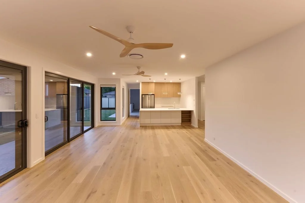 Open-concept living area with large windows and sliding glass doors, light wood flooring, and a modern kitchen with light wood cabinetry and white countertops.