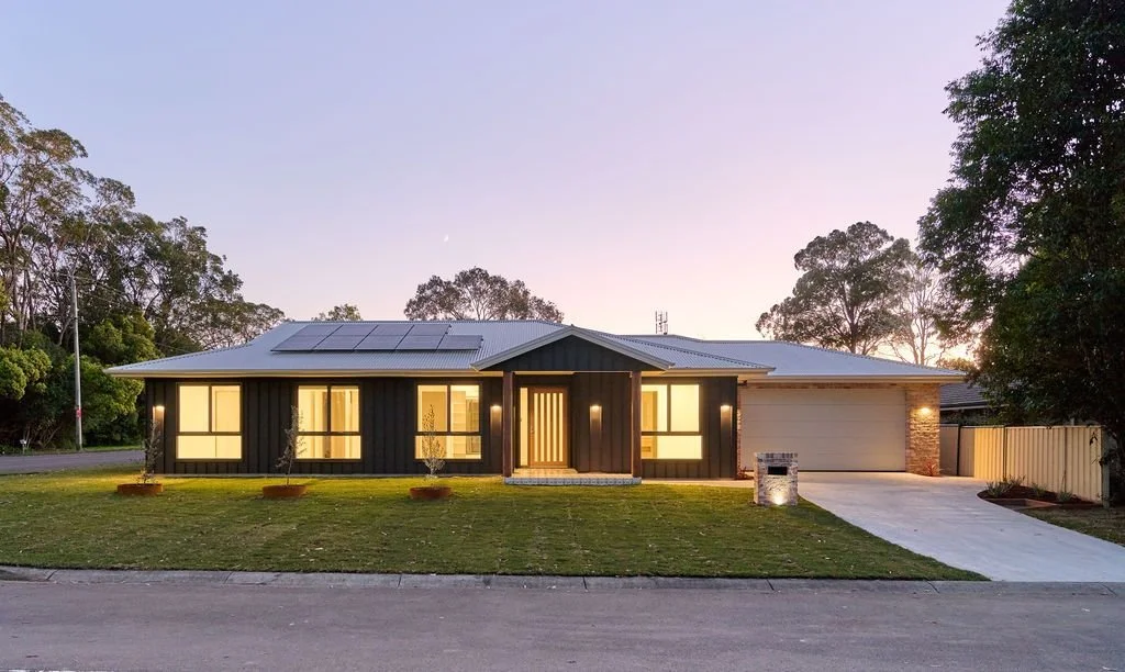 Modern single-story house with solar panels on the roof, illuminated windows, front yard, driveway, and a mailbox, surrounded by trees at sunset.