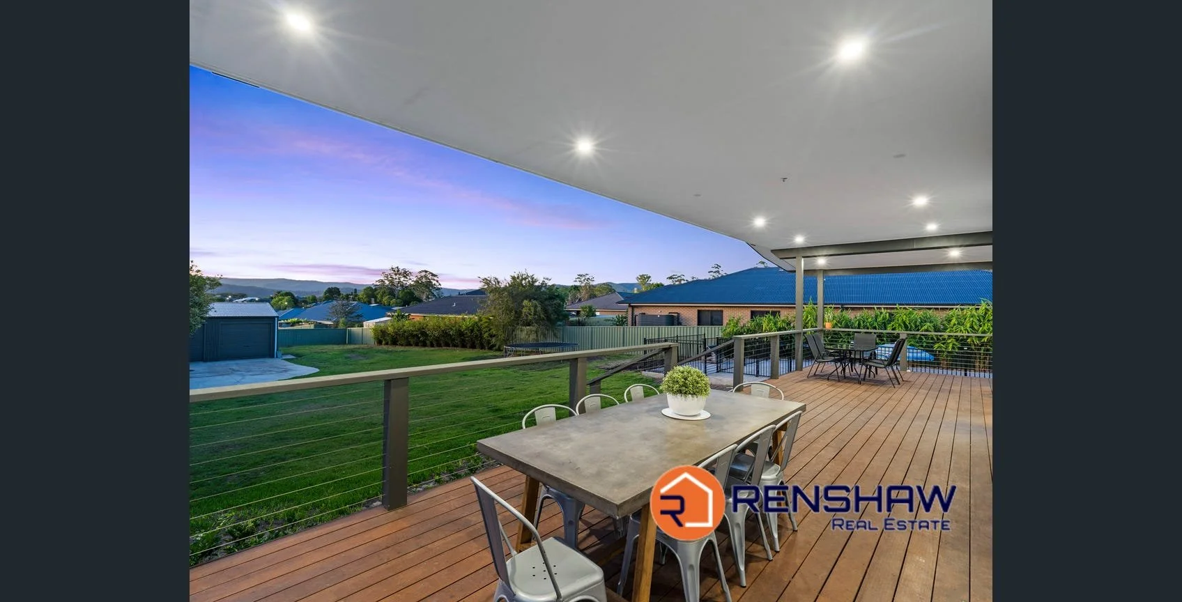 Modern outdoor balcony with wooden flooring, metal railing, a dining table with chairs and a potted plant, additional seating in the background, overlooking a green yard and neighboring houses with trees and hills in the distance at dusk.