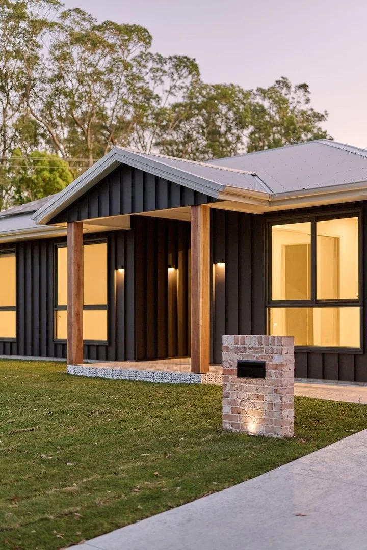 Modern house with black vertical siding, wooden porch pillars, and large windows illuminated from inside, set on a grassy yard with trees in the background.
