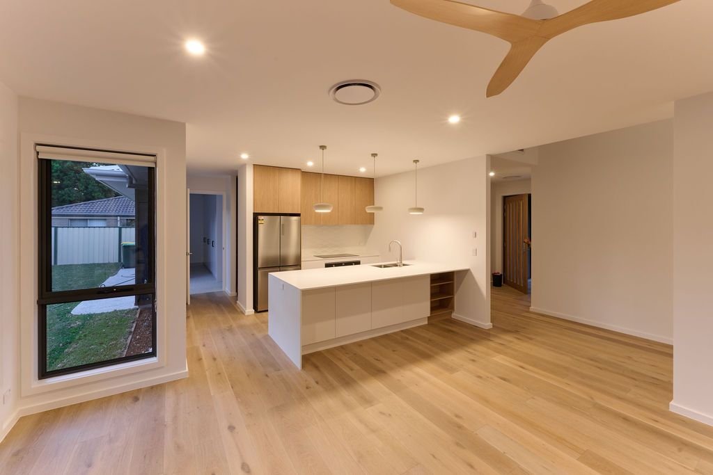 Open-concept kitchen and living area with hardwood floors, modern white island, stainless steel refrigerator, beige cabinetry, and pendant lighting, with large window overlooking a backyard.