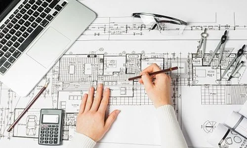 Person working on architectural blueprints with tools, a laptop, glasses, and a calculator on a desk.