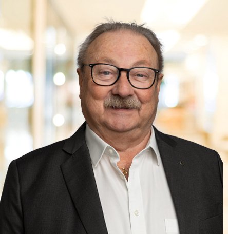 Ein älterer Mann mit Brille, schwarzen Haaren und einem Schnurrbart, trägt einen schwarzen Anzug und ein weißes Hemd, steht in einem hellen, modernen Innenraum.