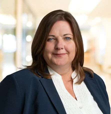 Eine Frau mit dunklen Haaren in einem blauen Blazer und weißem Hemd, die in einem modernen Gebäude steht.