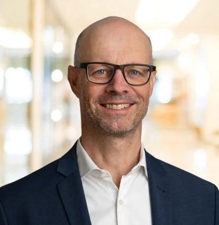 Ein Mann in einem dunklen Anzug mit Brille, lächelnd, im Innenraum eines Gebäudes.