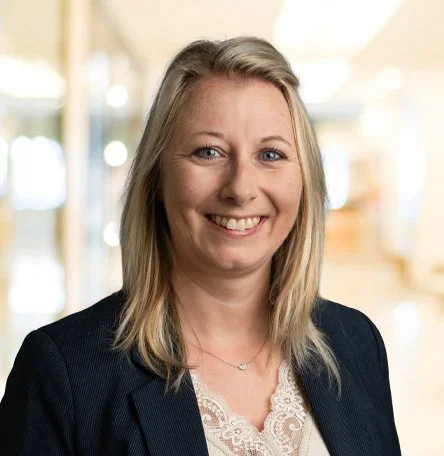 Ein Porträt einer lächelnden Frau mit blonden Haaren in einem dunklen Blazer vor einem unscharfen Bürohintergrund.