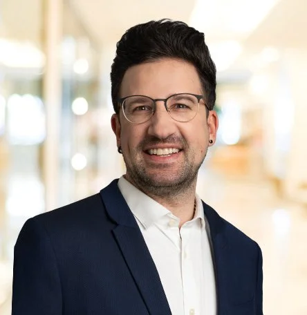 Porträt eines lächelnden Mannes mit dunklem Haar, Brille, Anzug und weißem Hemd vor unscharfem, hellem Hintergrund.