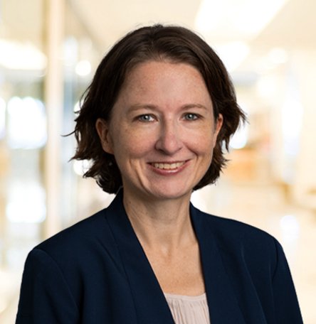 Porträt einer lächelnden Frau mit dunklen, lockigen Haaren, im beruflichen Umfeld, trägt dunkle Jacke und helles Oberteil.