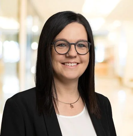Porträt einer lächelnden Frau mit schwarzen Haaren, Brille und schwarzem Blazer in einem hell erleuchteten Innenraum.