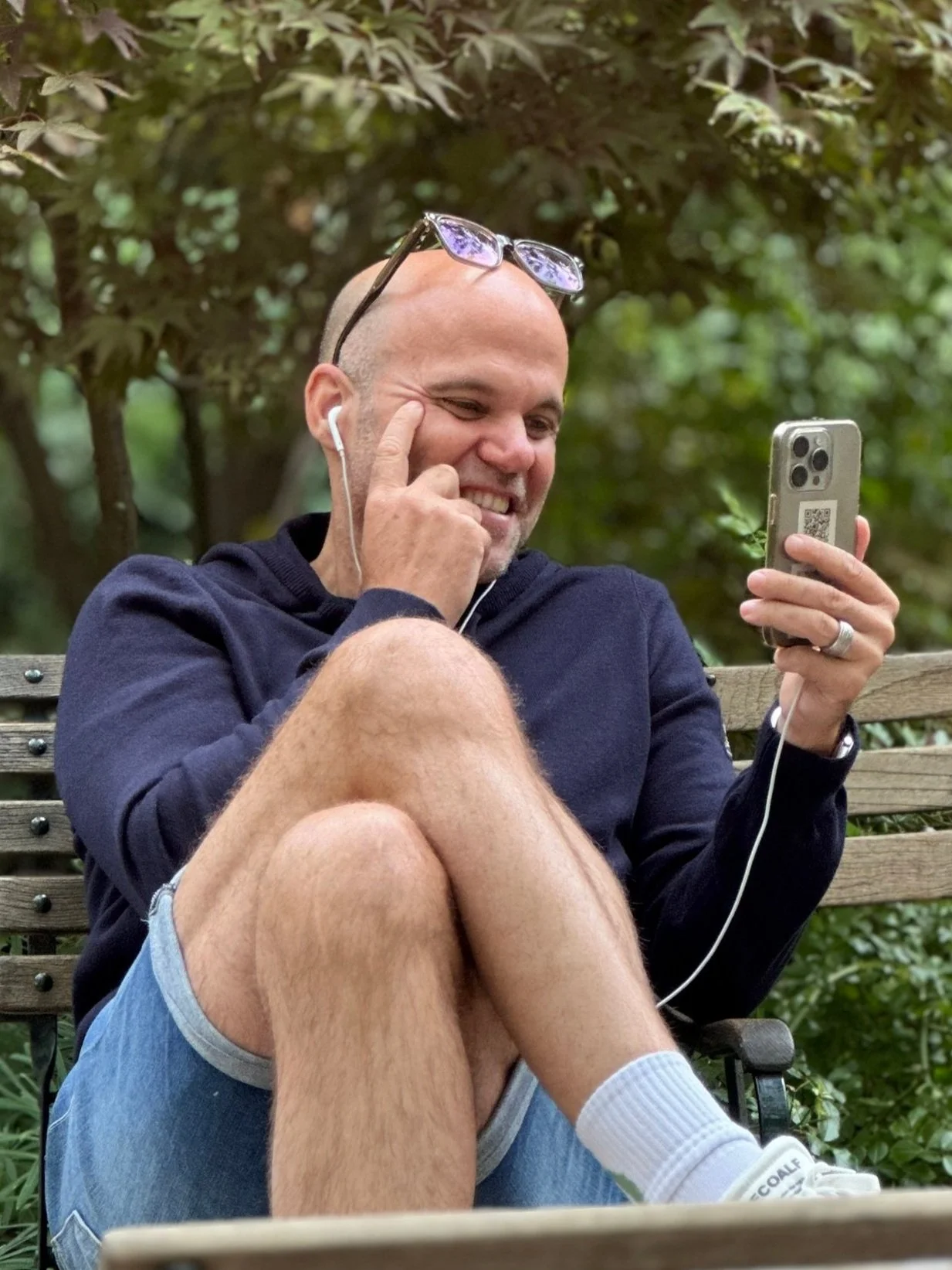 Ein lachender Mann sitzt auf einer Parkbank im Grünen, trägt eine schwarze Jacke, Shorts, weiße Socken und Schuhe, und benutzt sein Smartphone, während er eine Ohrhörer trägt.