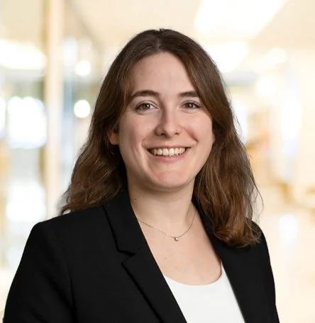 Porträt einer lachenden Frau mit braunen, lockigen Haaren, in einem schwarzen Blazer und weißen Top, im Büro.