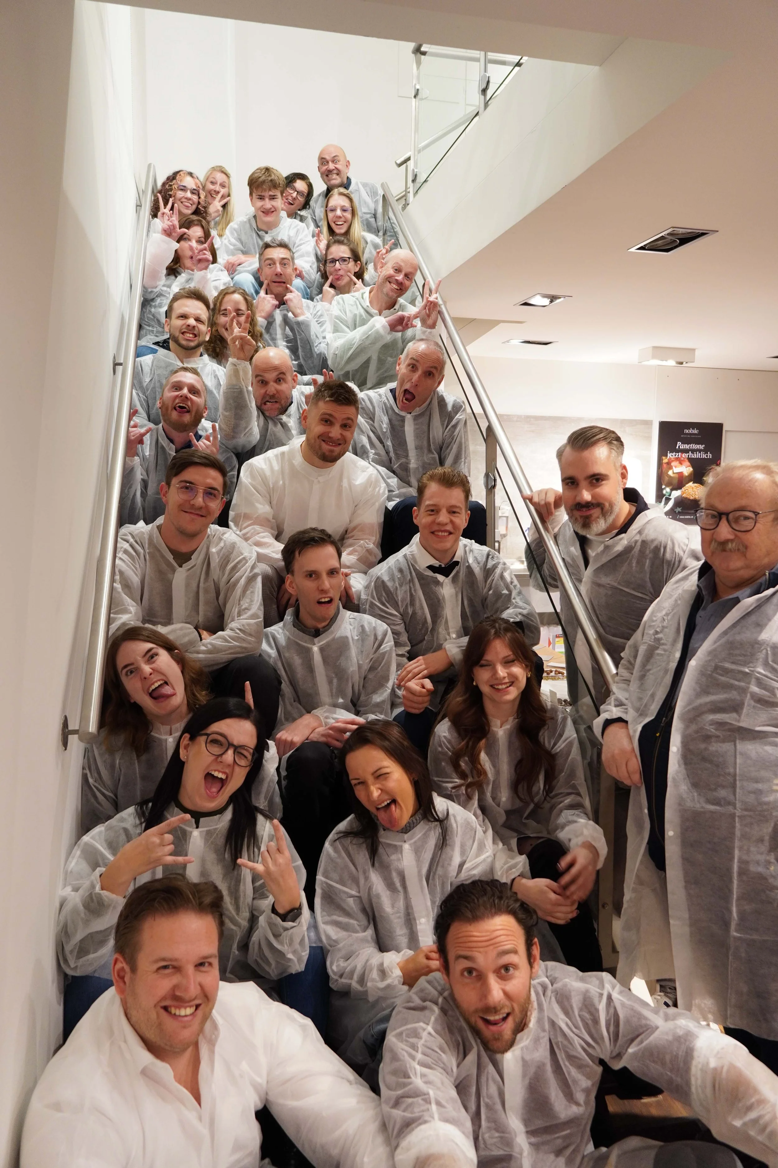 Eine große Gruppe von Menschen, die alle weiße Schutzkittel tragen, versammelt sich auf einer Treppe in einem Gebäude und posiert fröhlich für ein Gruppenfoto.