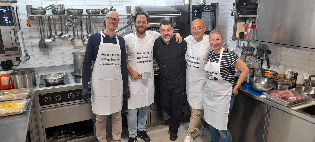 Fünf Personen in einer professionellen Küche, tragen Kochschürzen, lachen und stehen eng beieinander.