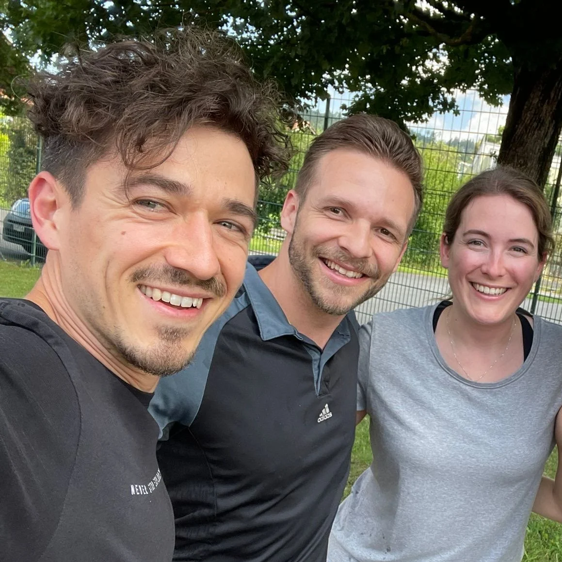 Drei lachende Menschen in einem Park, zwei Männer und eine Frau, die ein Selfie machen.