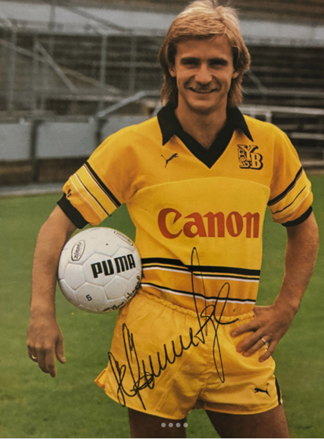 Der ehemalige Profi-Fussballspieler Roland Schönenberger im gelben YB-Trikot, hält einen weißen Puma-Fußball, steht auf einem Fußballfeld, lächelt.