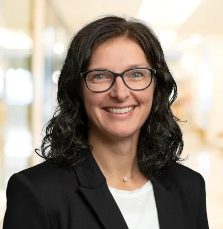 Porträt einer lächelnden Frau mit schwarzen, lockigen Haaren, Brille und Business-Kleidung in einem hellen Innenraum.