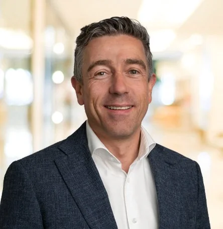 Ein lachen Mann in formeller Kleidung vor hell erleuchtetem Hintergrund.