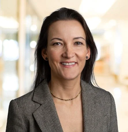 Ein professionelles Portrait einer Frau, die im Büro oder in einem geschäftlichen Umfeld steht.