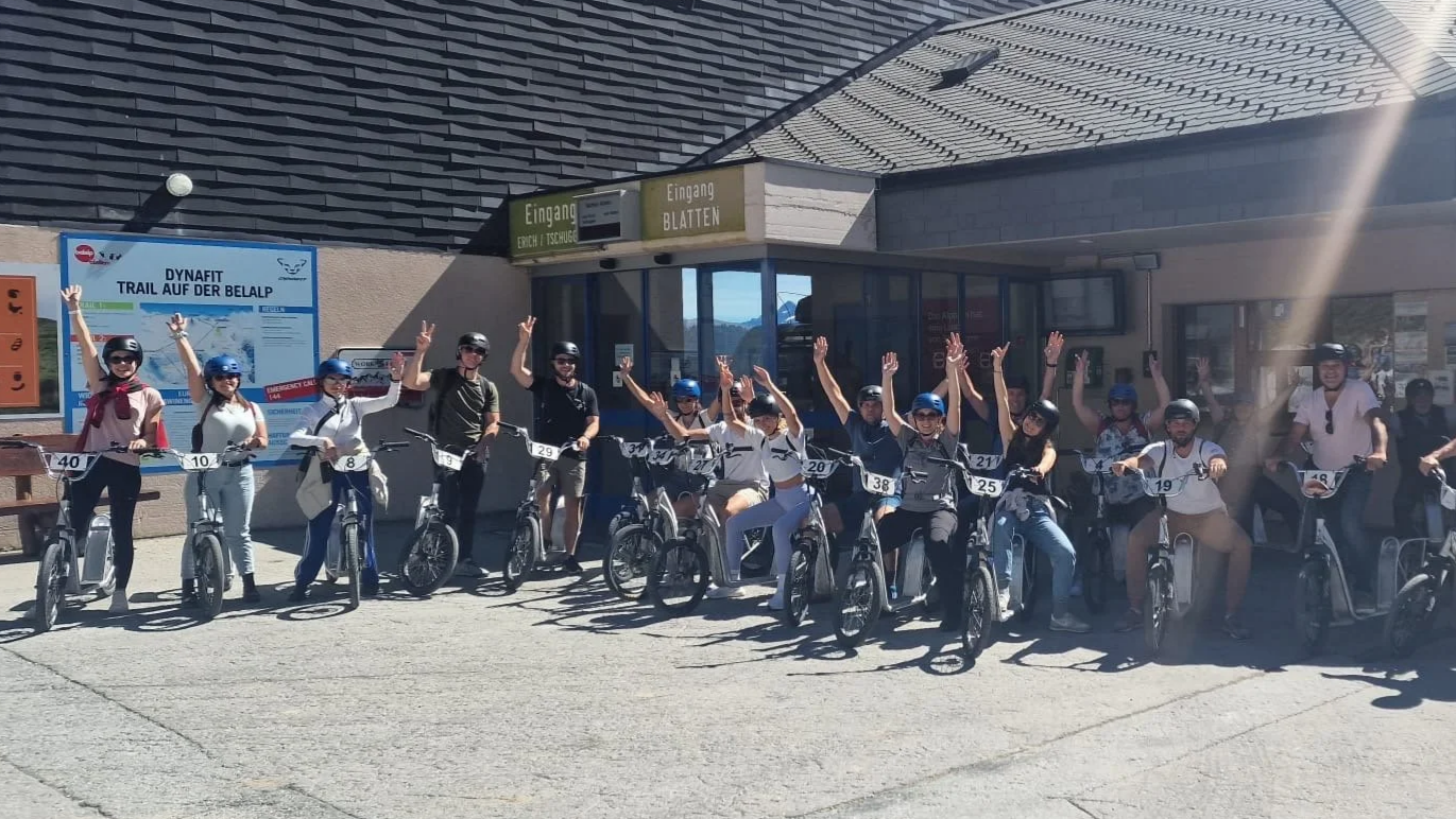 Gruppe von Personen mit Fahrrädern, alle tragen Helme, vor einem Gebäude mit einer Tür und Schildern, einige winken, es ist sonnig.