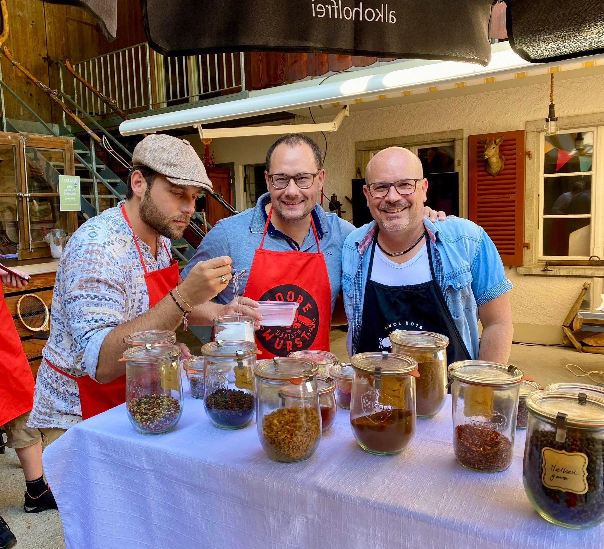 Drei Männer stehen hinter einer Tafel mit verschiedenen getrockneten Gewürzen oder Kräutern in Gläsern, beim Kochen oder Würzen, im Freien vor einem Haus.