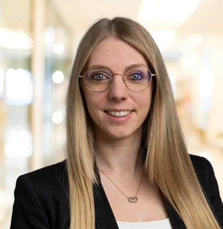 Porträt einer jungen Frau mit langen blonden Haaren, Brille und einem schwarzen Blazer, im Hintergrund eine helle, moderne Umgebung.