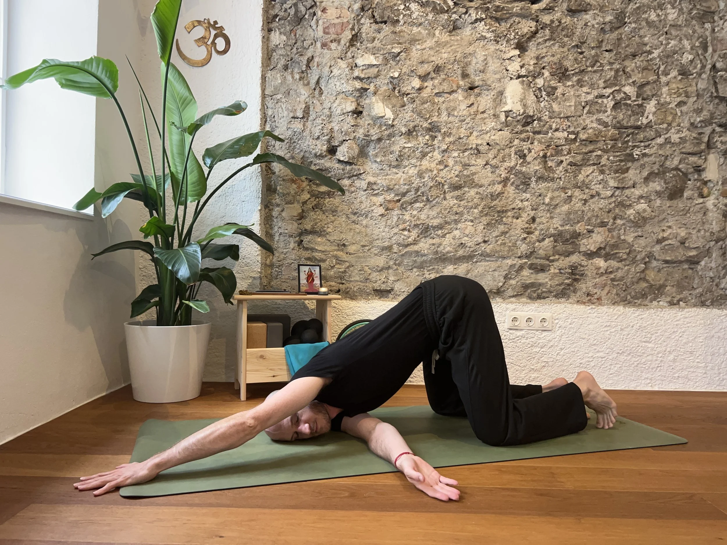 Gregor Eistert practicing yoga in a room with a brick wall, a large green plant, and a small wooden shelf with a framed picture and candles, doing a twisted puppy pose.