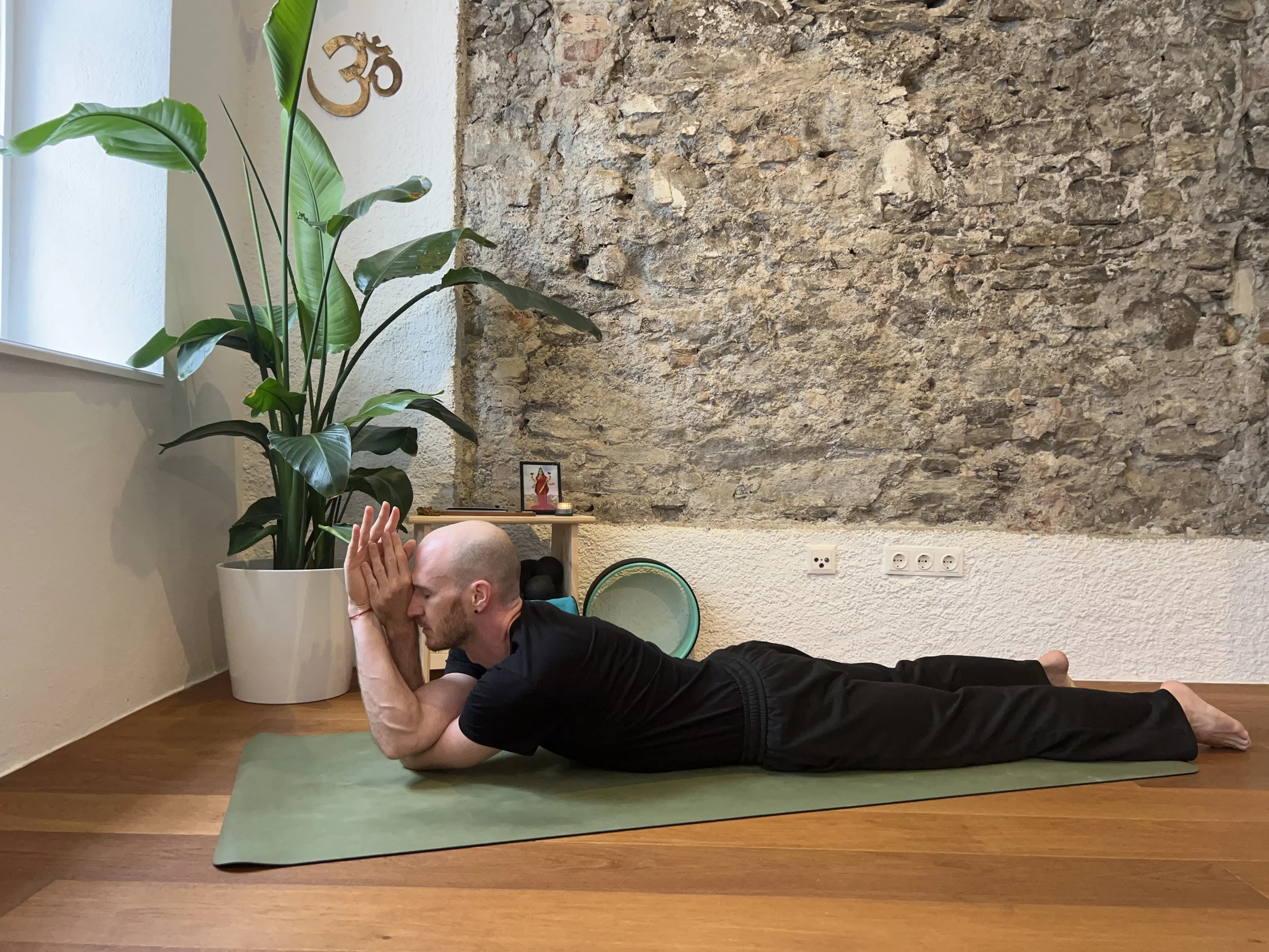 Gregor Eistert practicing sphinx with eagle arms in a room with a large brick wall, a potted plant, and a small altar with a picture and a candle. He is lying on a green yoga mat, with his head resting on his hands, and his eyes closed.
